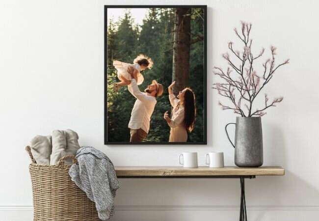 A canvas print hanging in a white modern entryway. The print is of a husband playing with his daughter and the mom looking on with love. 